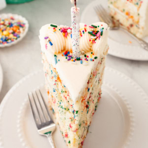 Piece of birthday cake with sprinkles with a candle. A fluffy homemade Funfetti birthday cake with rainbow sprinkles topped with a light cream cheese buttercream frosting and an array of sprinkles. This Confetti Cake recipe is the perfect birthday cake!