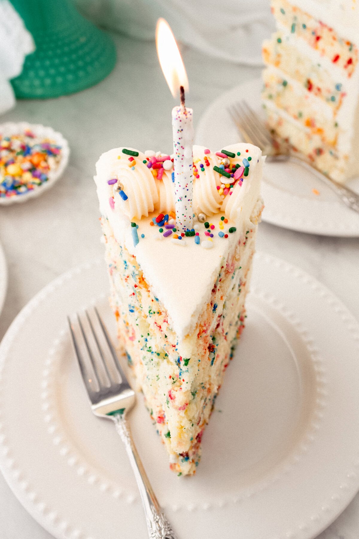 Piece of birthday cake with sprinkles with a candle. A fluffy homemade Funfetti birthday cake with rainbow sprinkles topped with a light cream cheese buttercream frosting and an array of sprinkles. This Confetti Cake recipe is the perfect birthday cake!